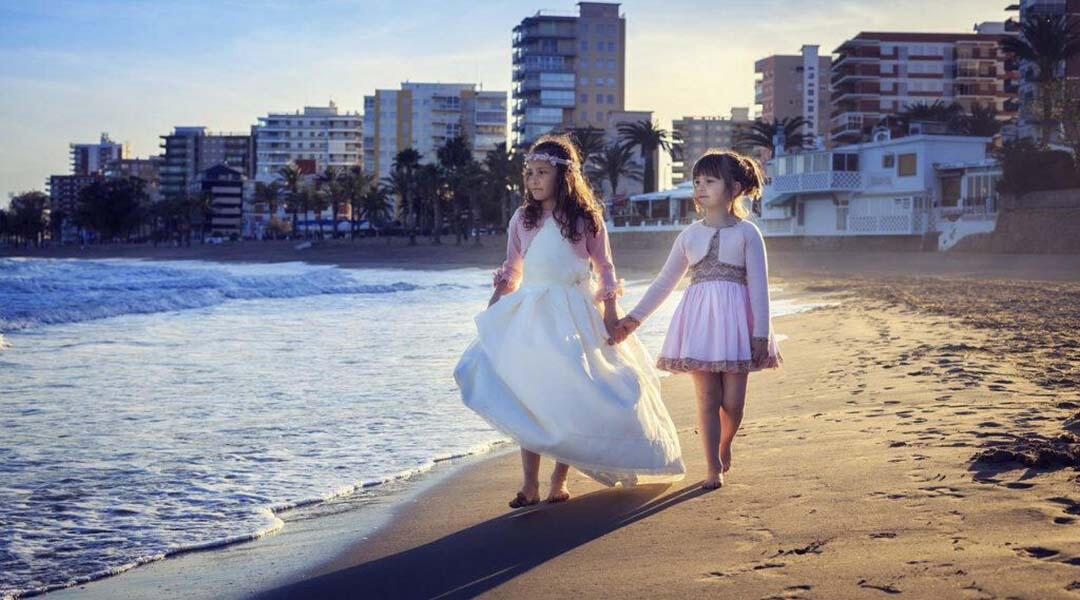 Niña vestida de comunión pasea por la playa con su hermana de la mano.